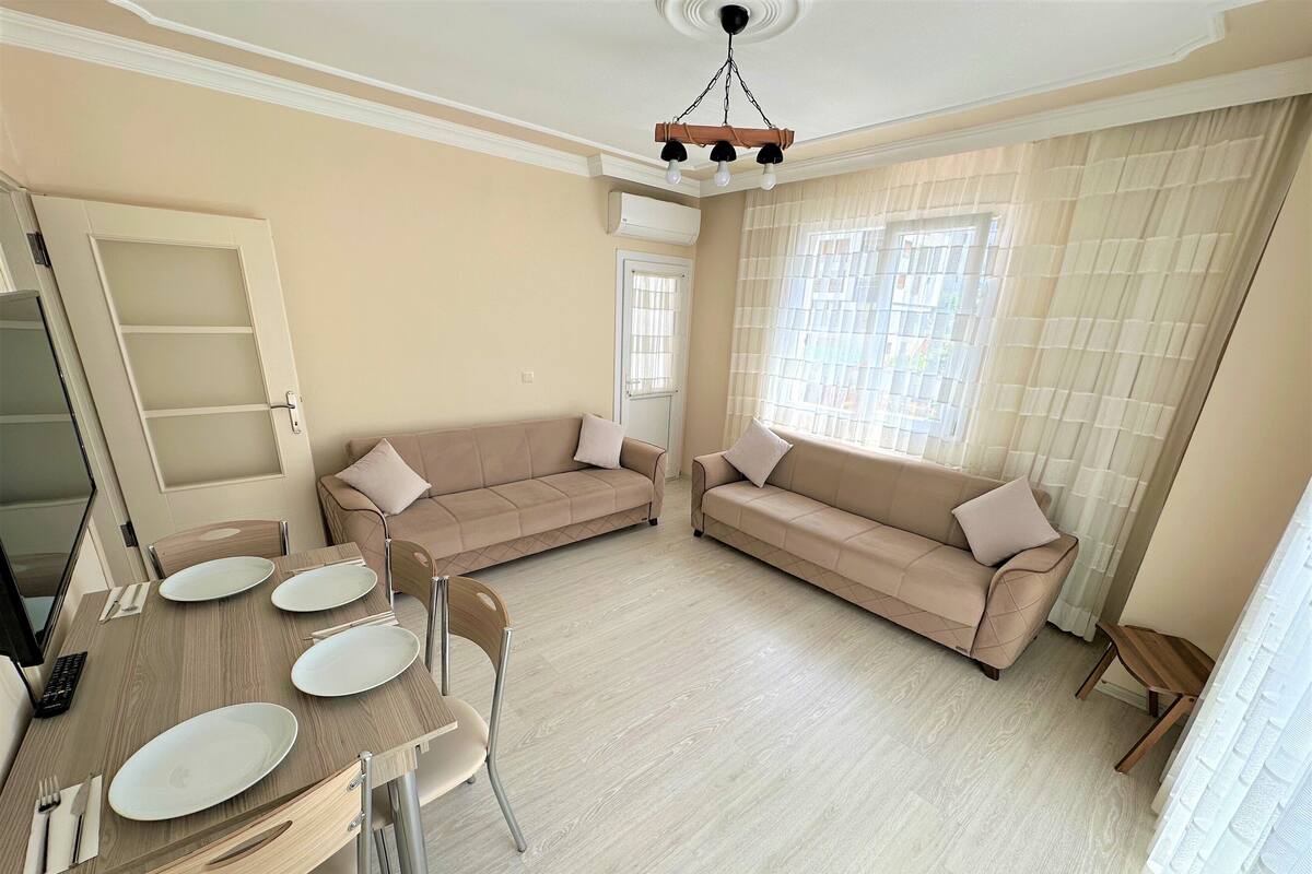 A living room with a beige sofa, a dining table with plates, and a ceiling light fixture.