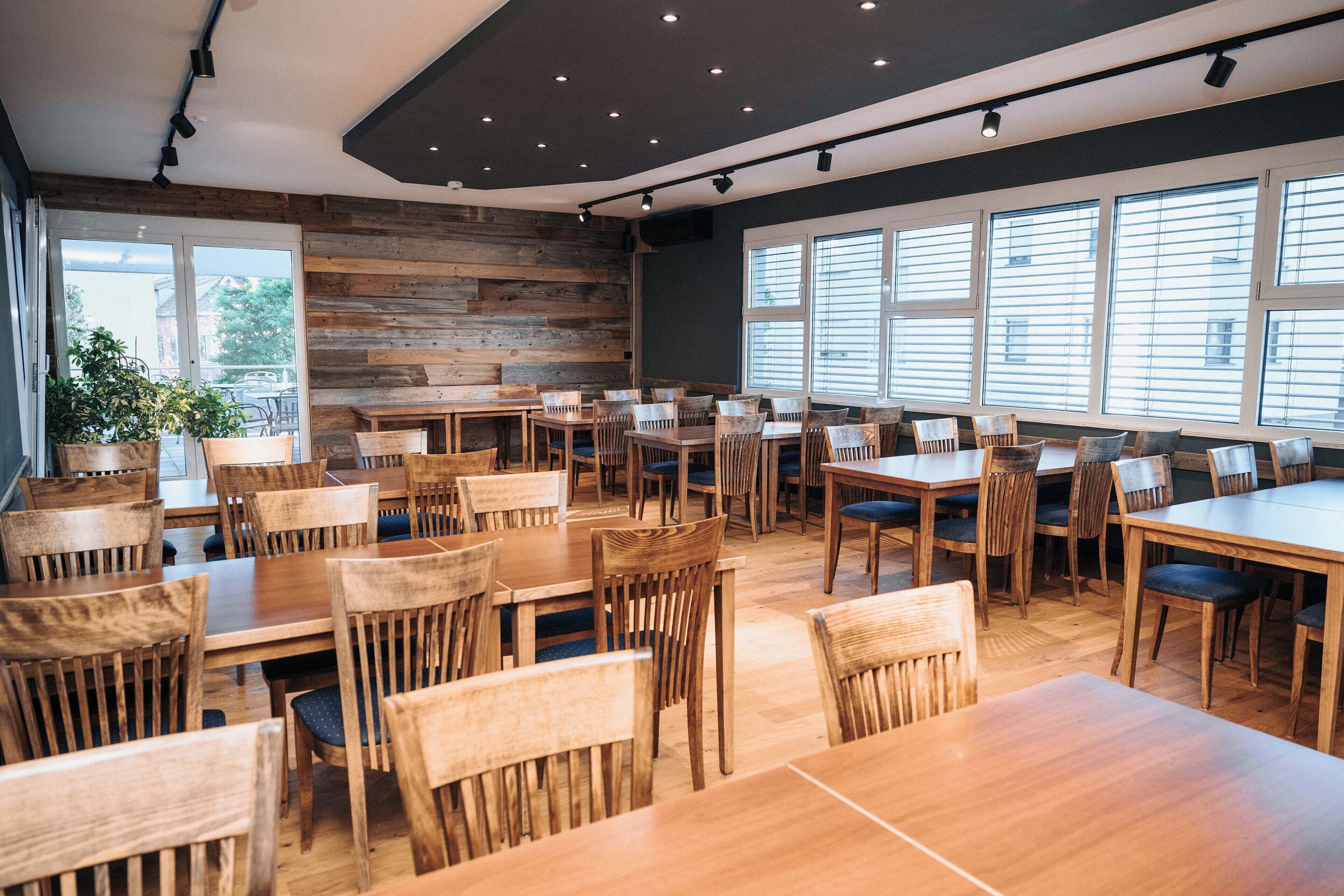 A restaurant with multiple wooden tables and chairs, large windows with blinds, and a wooden wall.