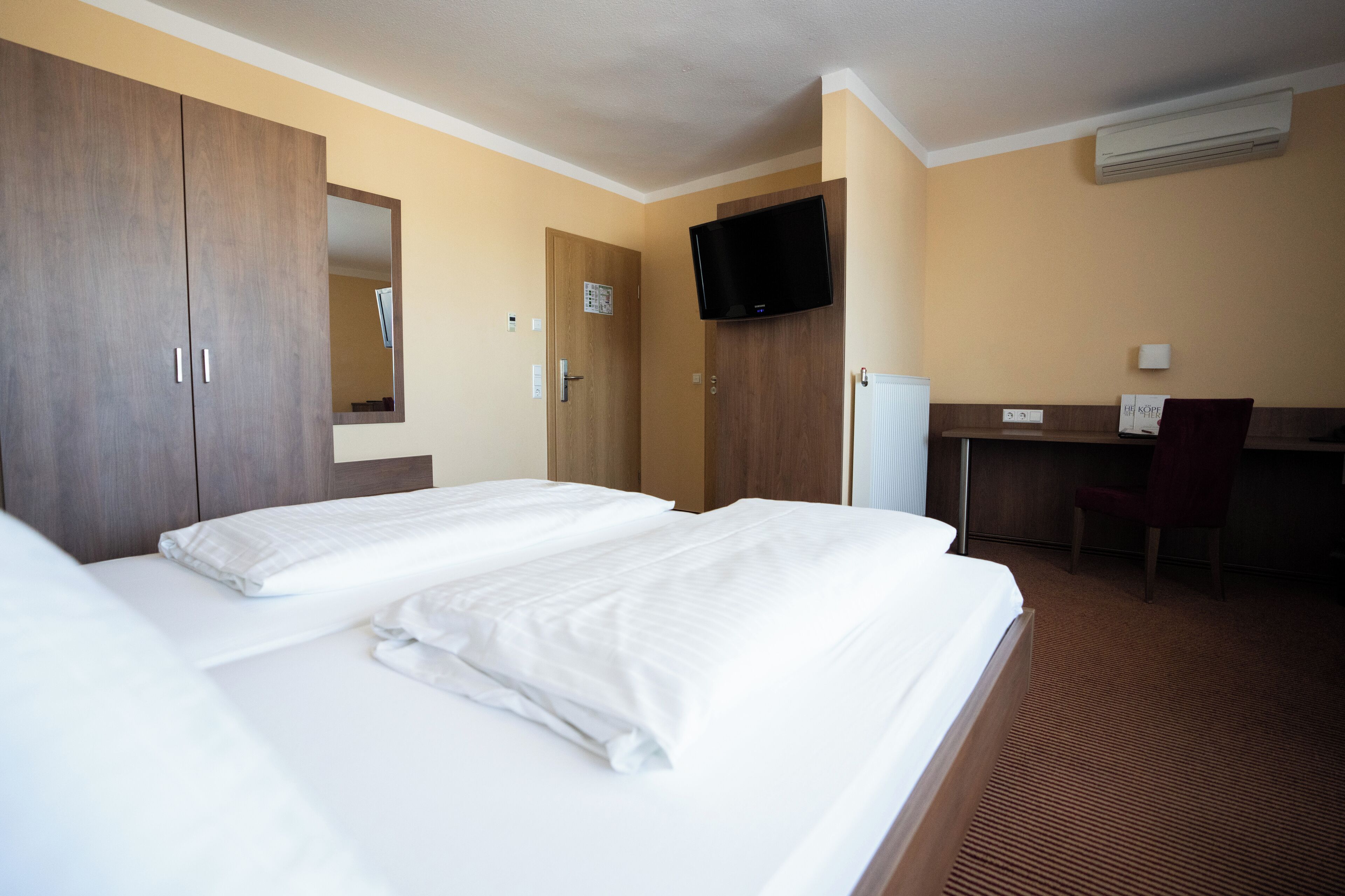 A hotel room with two beds, a desk, a chair, a television, and an air conditioning unit.