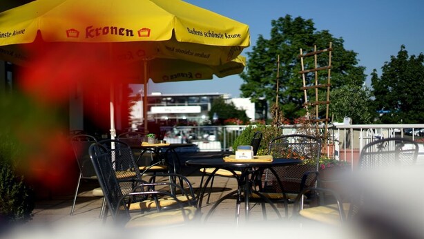 An outdoor seating area with tables and chairs under a yellow umbrella, with a building and trees in the background. - Europa Hotel (Kehl)
