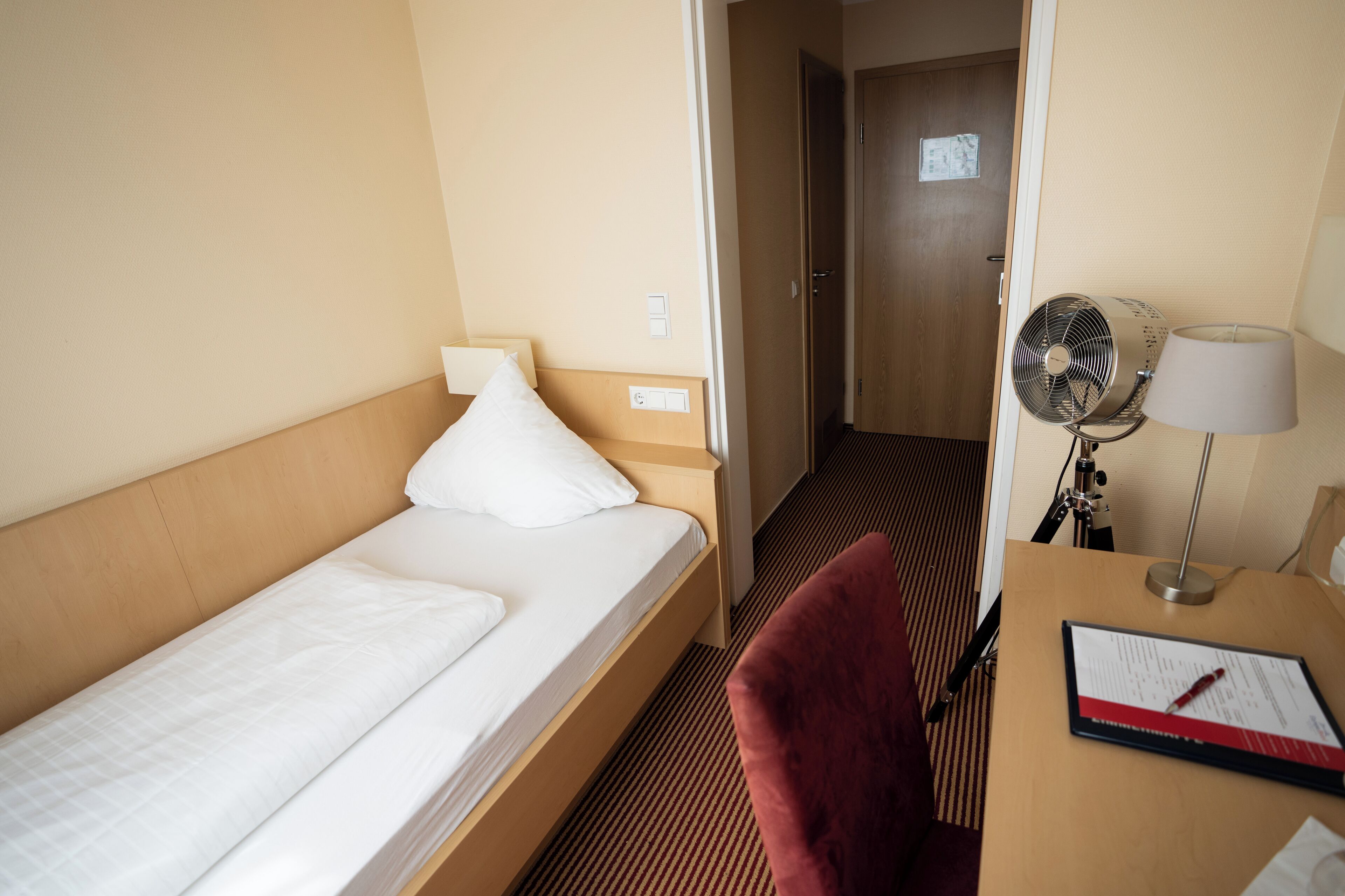 Habitación de hotel con una cama individual, un escritorio con lámpara, una silla y un ventilador de pie.