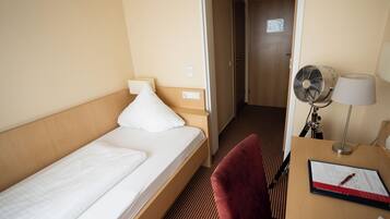 Habitación de hotel con una cama individual, un escritorio con lámpara, una silla y un ventilador de pie.