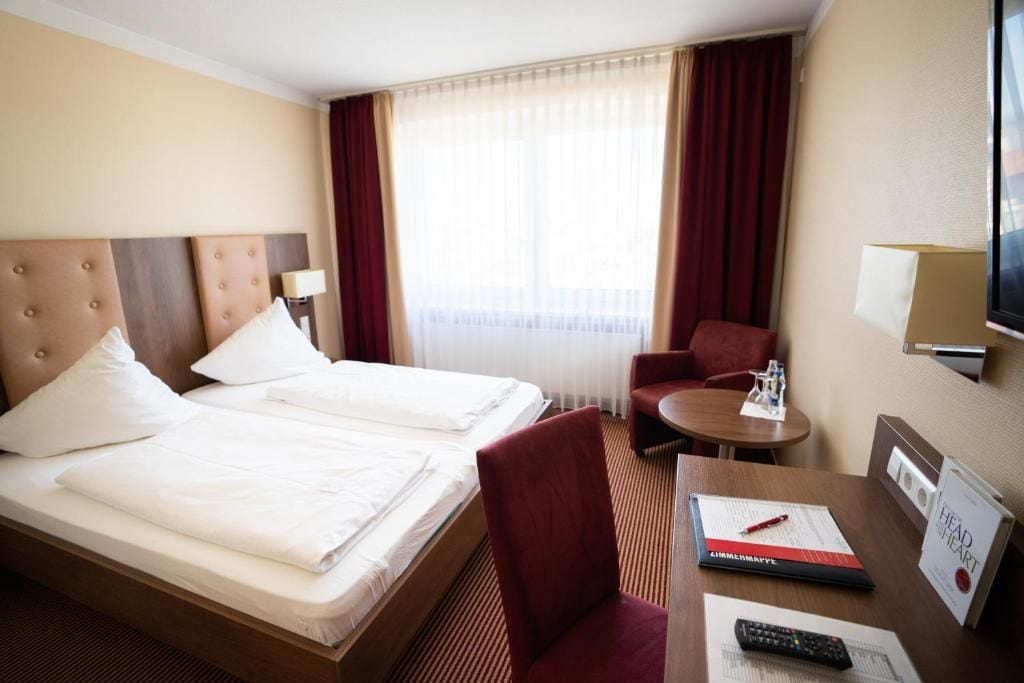 A hotel room with two beds, a desk with a remote control, a chair, a small table with a bottle, and a television on the wall.