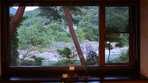 Een traditionele Japanse kamer met tatami vloeren, een lage tafel met een hete pot en diverse gerechten, en een raam met uitzicht op een bebost gebied.