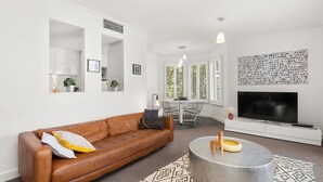 A modern living room with a brown leather sofa, a round coffee table, and a TV on a white stand. There are two pendant lights, a dining table with chairs, and a large window with blinds. - BOUTIQUE STAYS - Wellington Mews (East Melbourne)