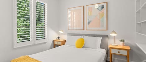 Un dormitorio con cama, dos mesitas de noche, un tocador, una ventana con persianas y dos obras de arte enmarcadas en la pared.