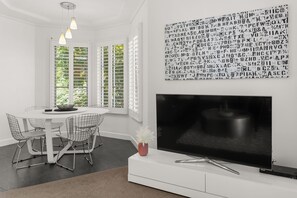A modern living room with a white TV stand, a flat-screen TV, a dining area with wireframe chairs, and a wall with a black and white pattern.