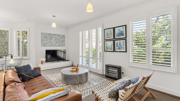 A modern living room with a leather sofa, a round coffee table, and a fireplace. There are large windows with shutters and framed cityscape pictures on the wall. - BOUTIQUE STAYS - Wellington Mews (East Melbourne)