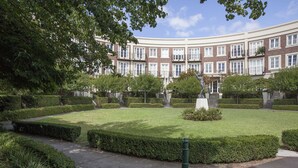 En velholdt gårdsplads med velplejede hække, en statue og en bygning med ensartede vinduer i baggrunden.