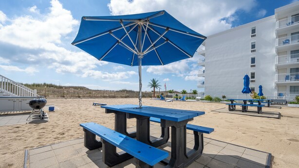 A beachside patio with a blue umbrella, picnic table, and barbecue grill.