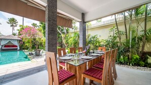 A dining area with a glass table and wooden chairs, surrounded by lush greenery and a pool. - Lush Private Pool | Shaded Gazebo | Seminyak (Seminyak)
