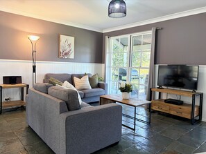A modern living room with a grey sofa, a coffee table, a TV on a wooden stand, and a large window with a view of the outdoors.