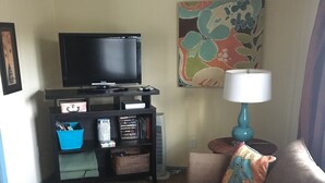 Una sala de estar con televisor de pantalla plana sobre un mueble bajo, una lámpara azul y un sofá con cojines de colores.
