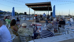 Un área de comedor al aire libre con sillas y mesas de metal, un escenario con músicos y vistas a barcos al fondo.