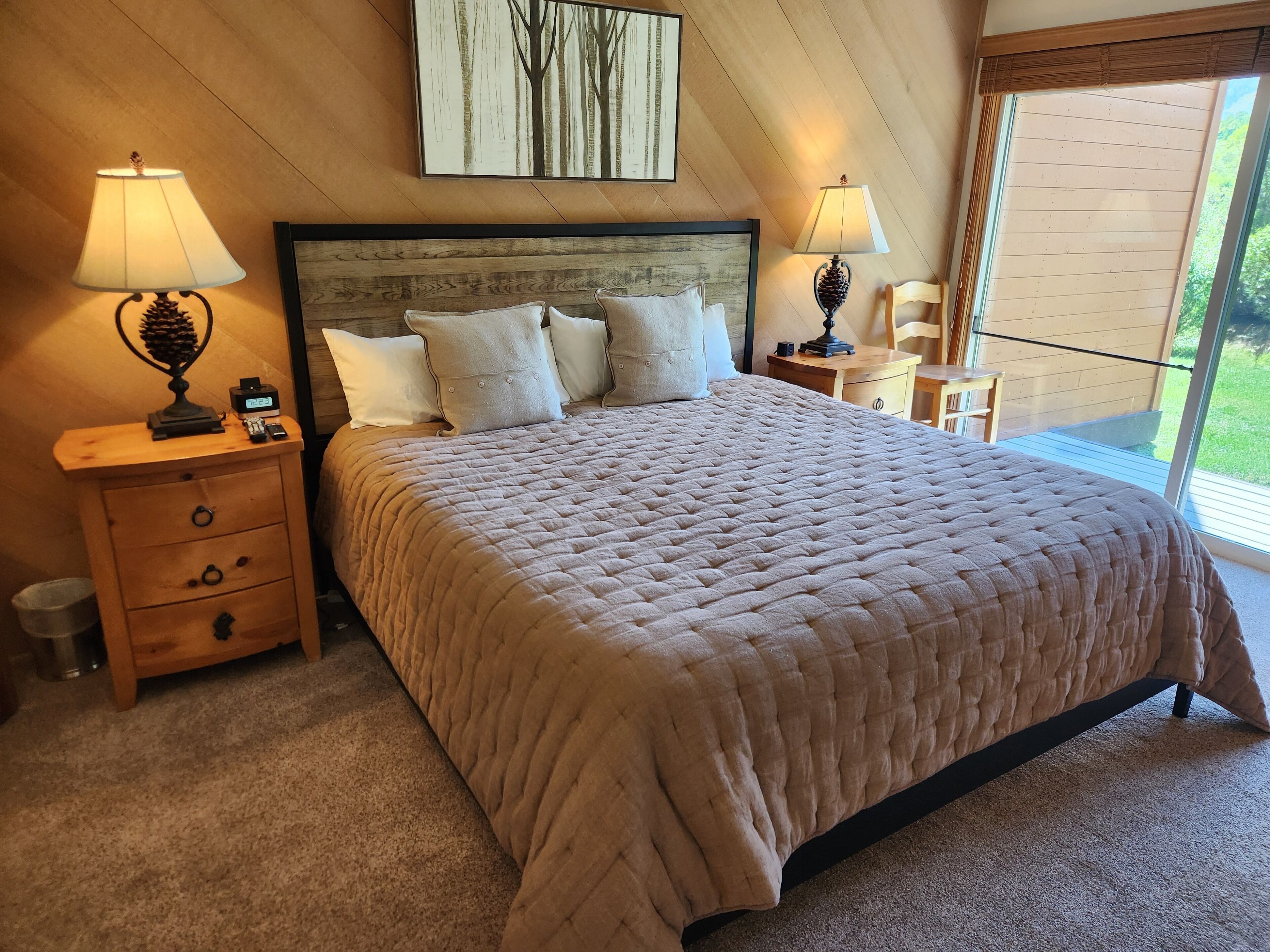 Een slaapkamer met een groot bed, twee nachtkastjes met lampen, een houten hoofdbord en uitzicht op het omliggende landschap door een groot raam.