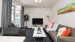 Una sala de estar moderna con un televisor de pantalla plana, una mesa de centro con libros, un sofá gris con almohadas naranjas y un ventanal con cortinas grises.