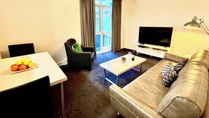 Una sala de estar con un sofá, televisión, una mesa de centro y una silla. Hay una ventana con cortinas y un equipo de aire acondicionado en la pared.