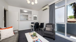 Una sala de estar moderna con un sofá gris, una mesa de centro blanca con revistas, un sillón gris, un comedor con una mesa blanca y sillas negras, y un ventanal con cortinas grises.