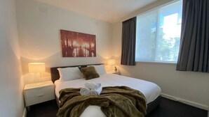 Habitación de hotel con una cama grande, una mesita de noche, una lámpara, un cuadro en la pared y una ventana con cortinas.
