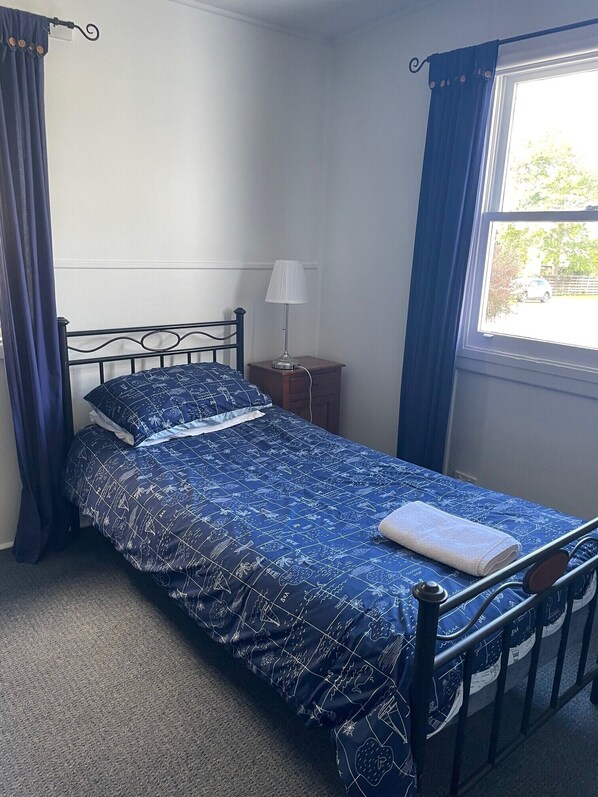 Un lit simple avec une couette à motifs, une table de chevet avec une lampe et une fenêtre avec des rideaux.