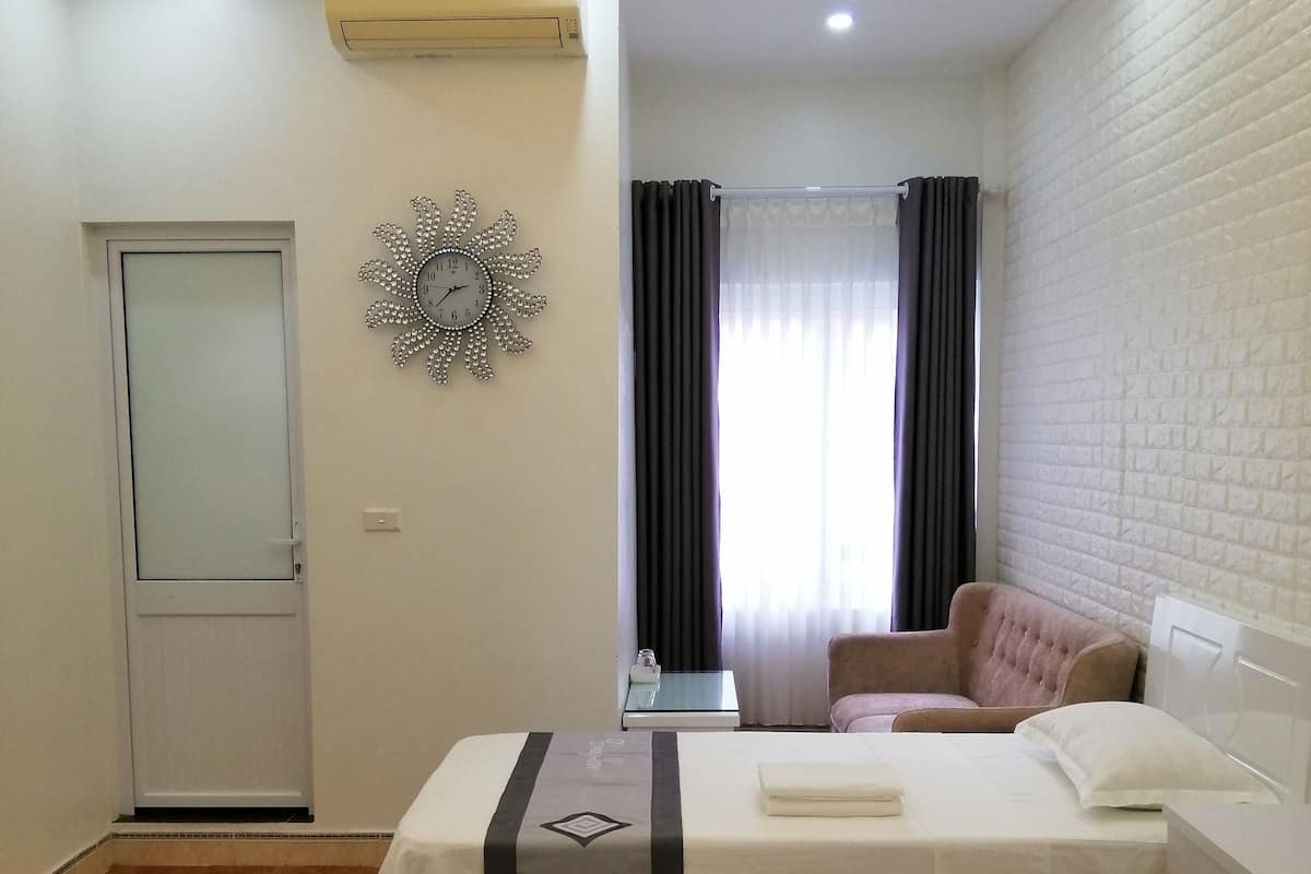 A hotel room with two beds, a ceiling fan, an air conditioning unit, a decorative clock, a window with curtains, and a brick-patterned wall.