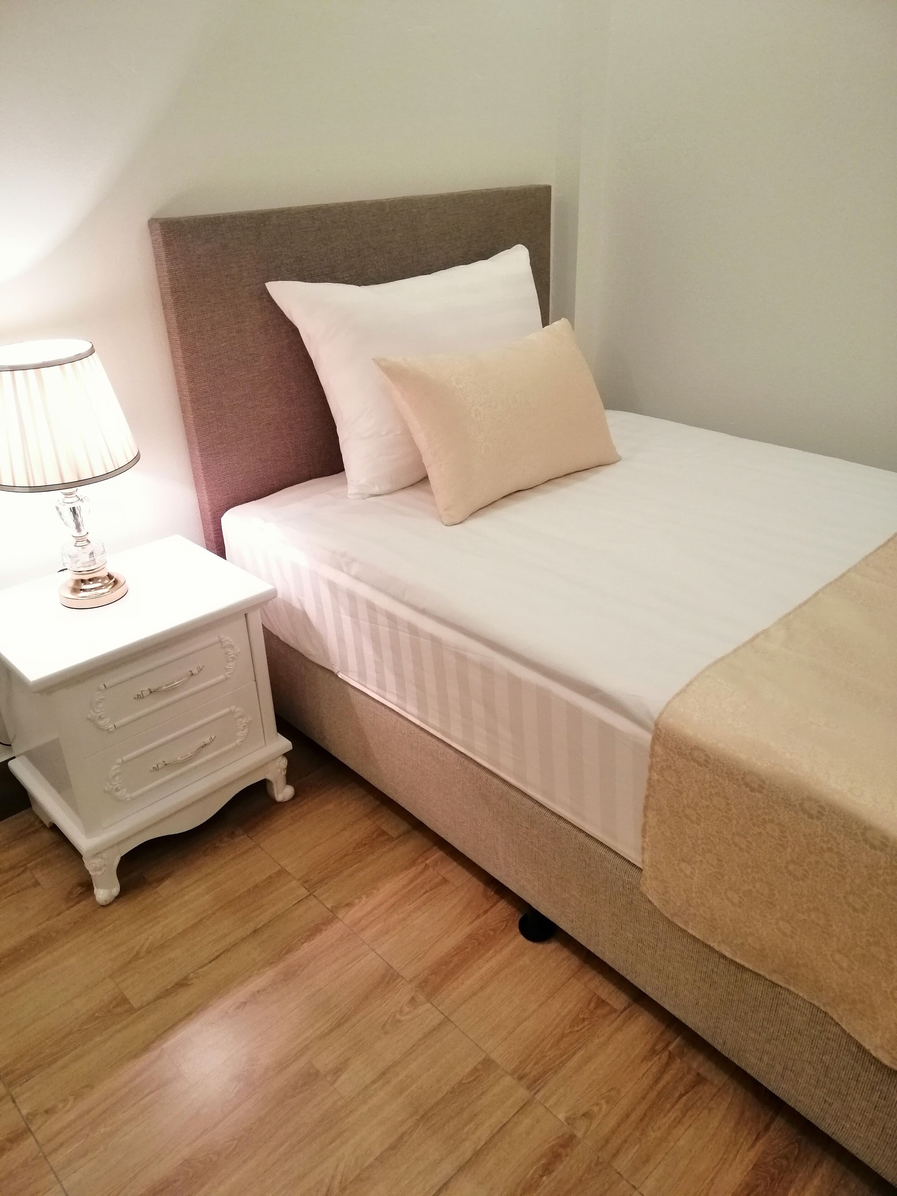 Una cama bien hecha con cabecera, mesita de noche con cajones y una lámpara.