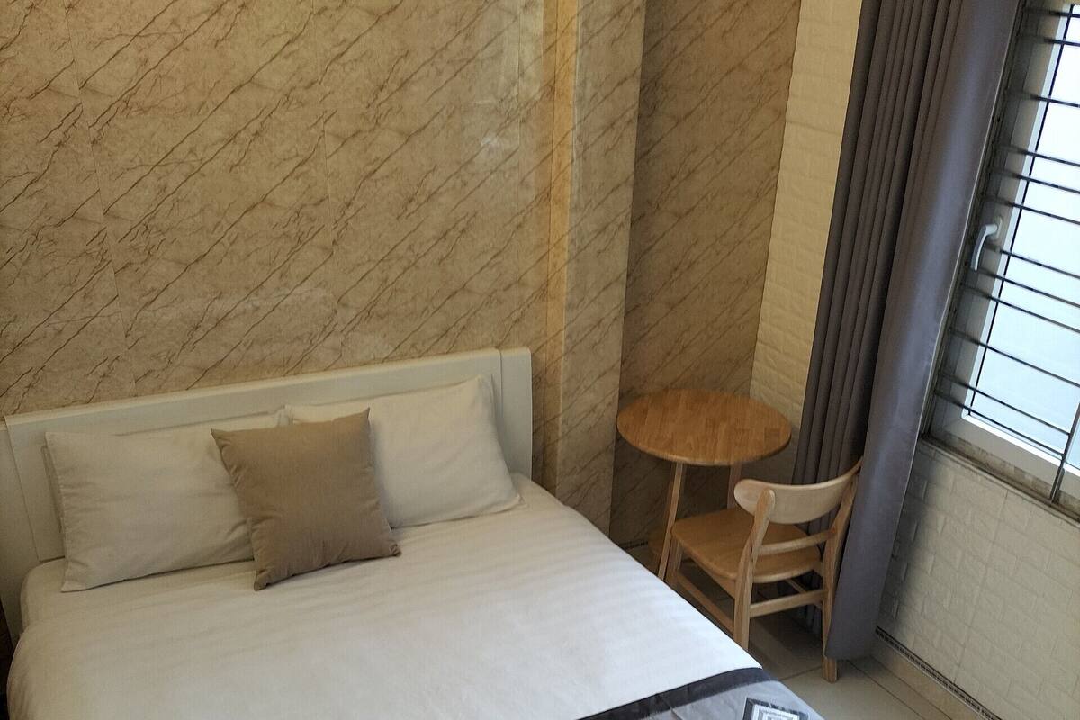 A neatly made bed with a grey and white bedspread, a small round table with a chair, and a window with curtains.