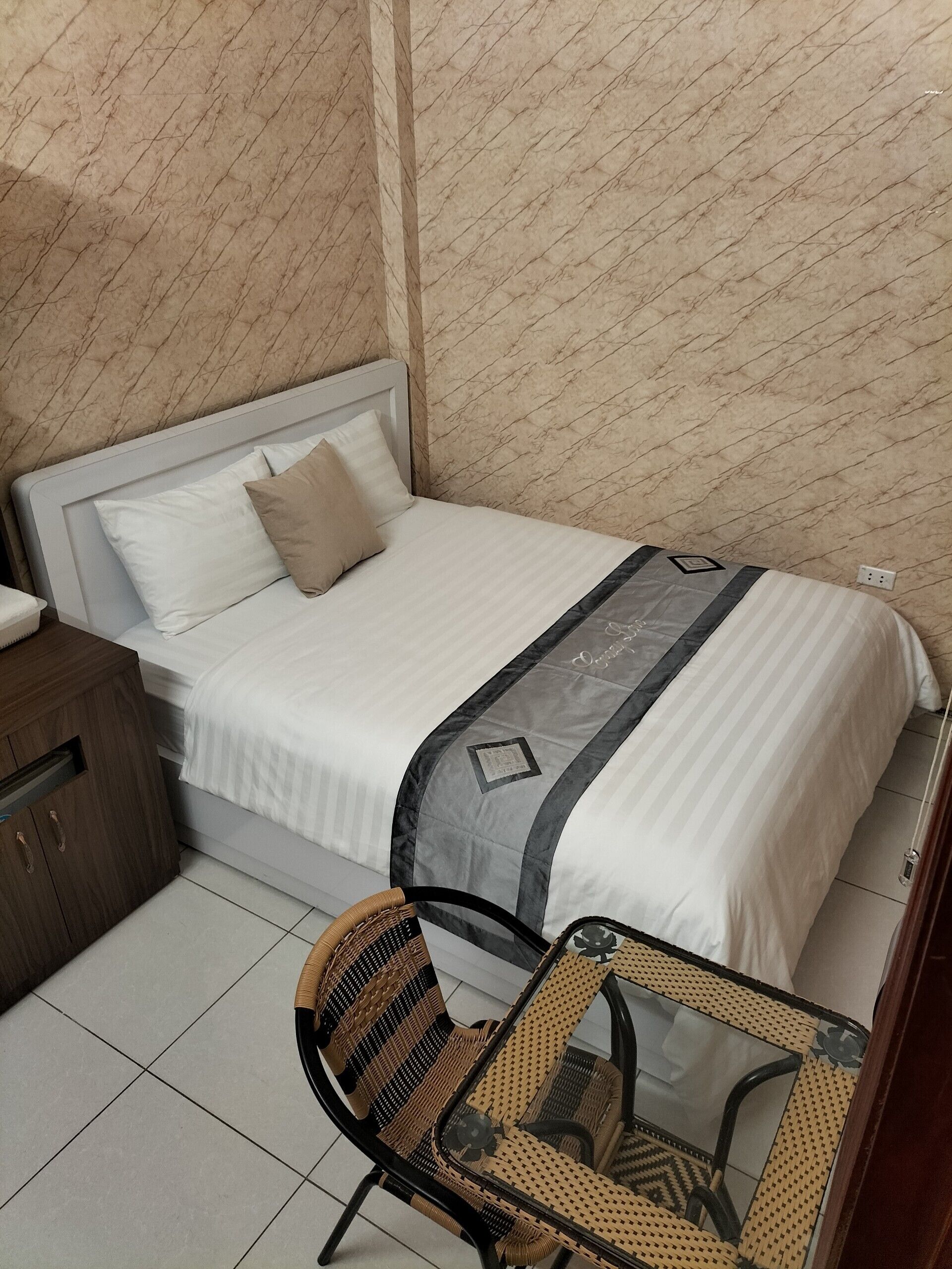 Una cama bien hecha con una manta a rayas, una mesita de noche de madera y una silla de mimbre con una mesa de vidrio.