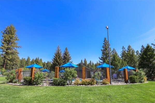 A well-maintained outdoor area with a pool, surrounded by a fence and blue umbrellas, set against a backdrop of tall trees.