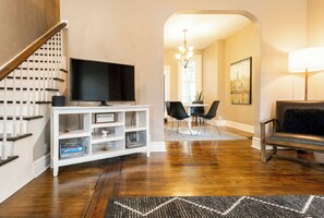 A living room with a flat-screen TV on a white entertainment unit, a dining area with a chandelier, and a black leather chair.