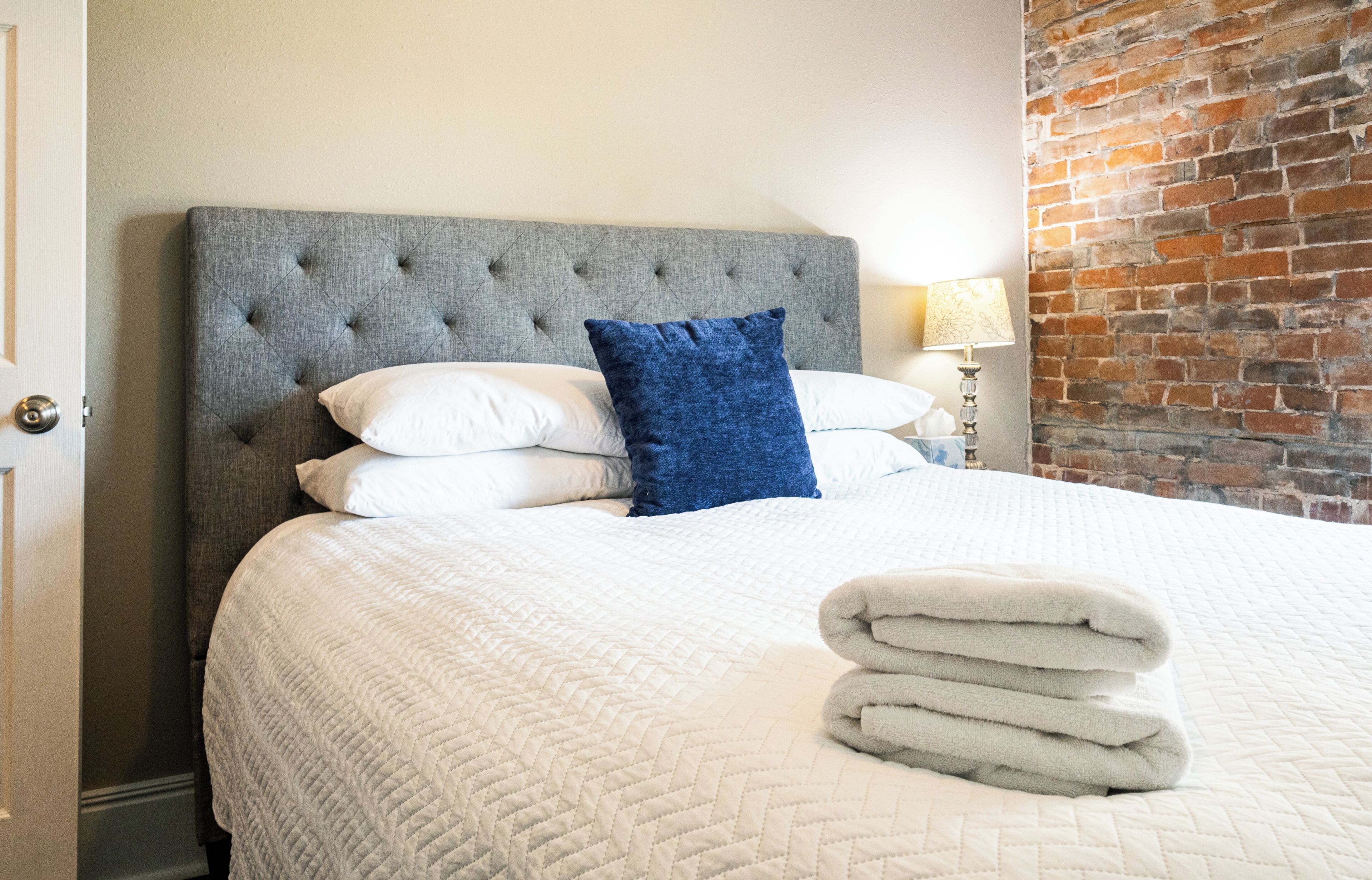 A hotel room with a large bed, a grey upholstered headboard, white bedding, a blue accent pillow, and a brick wall.