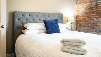 A hotel room with a large bed, a grey upholstered headboard, white bedding, a blue accent pillow, and a brick wall.