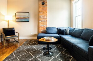 A modern living room with a sectional sofa, a small round table, a chair, a lamp, and a painting on the wall.