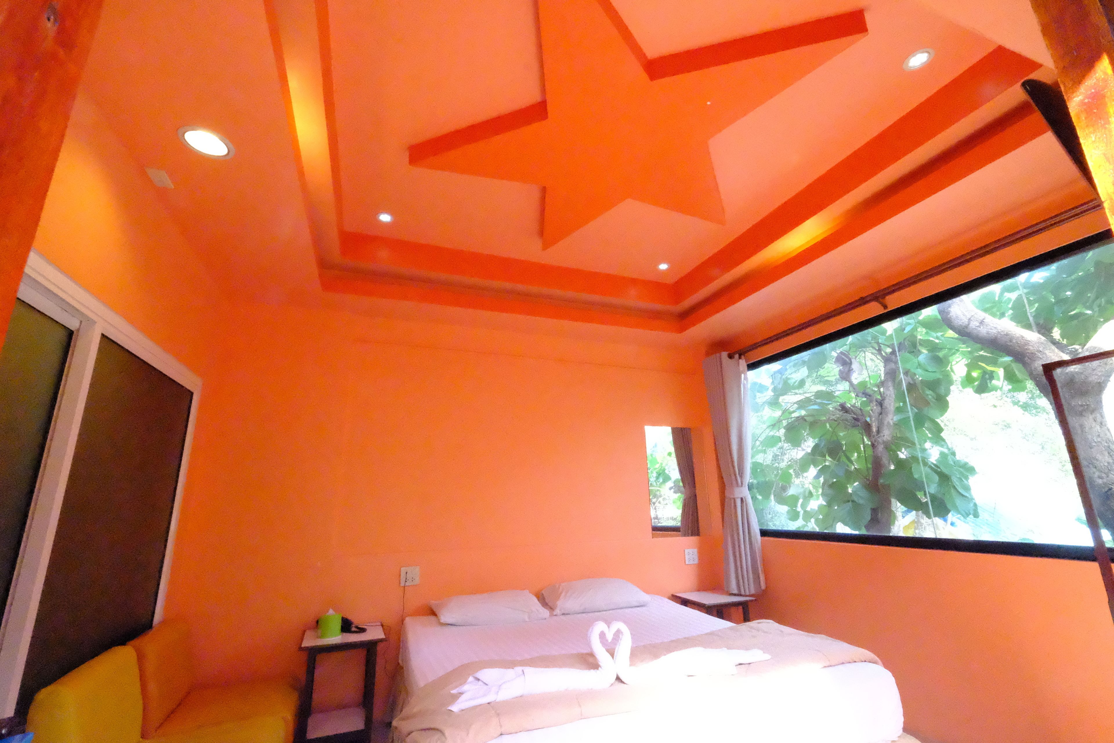 Una habitación con paredes naranja, una cama con sábanas blancas, una mesita de noche pequeña y una ventana con vistas a árboles.