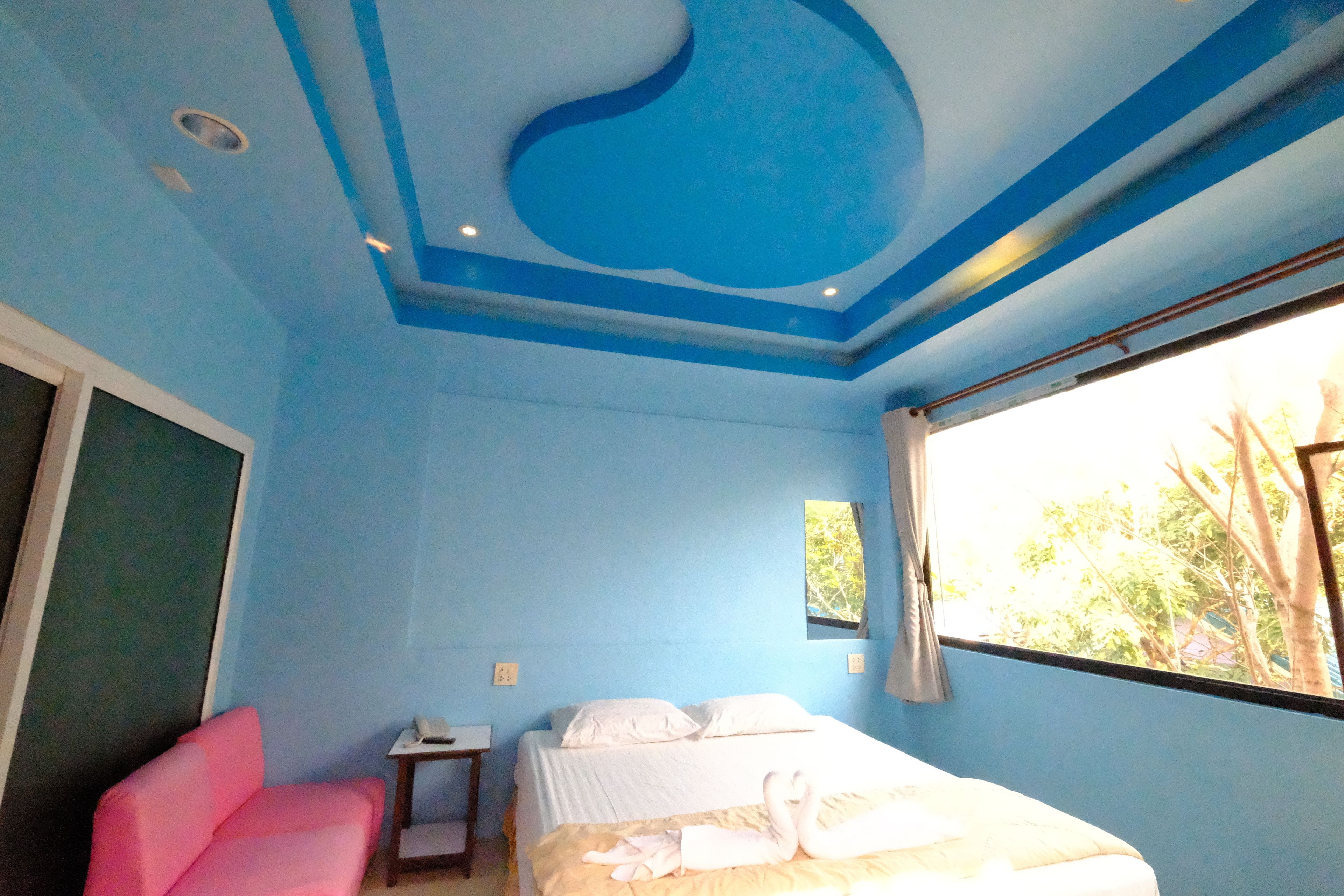 A room with a bed, a pink chair, a small table, and a window with a view of trees.