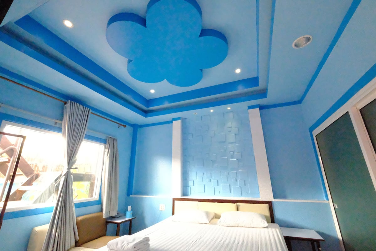 A hotel room with a bed, a nightstand, a window with curtains, a wall-mounted light fixture, and a ceiling with a floral design.