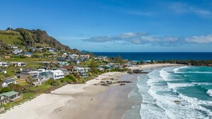 Un quartier résidentiel situé sur la côte, avec des maisons le long de la plage, une vue dégagée sur l’océan et un paysage vallonné en arrière-plan.