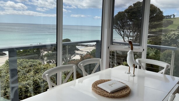 Une table blanche, agrémentée d’un set de table en fibres tressées et d’un carnet, est placée devant une vue panoramique sur l’océan, depuis un balcon où se trouvent des chaises blanches.