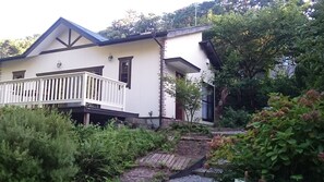 Una casa de dos pisos con balcón, rodeada de abundante vegetación y un pequeño jardín.