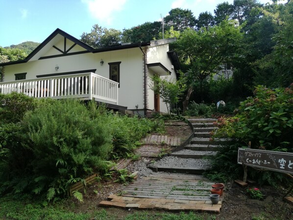 Una casa con fachada blanca y terraza de madera. Cuenta con un jardín con diversas plantas y un camino de grava que lleva a la casa.