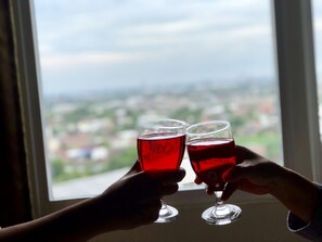 Two hands holding wine glasses with a city view through a window.