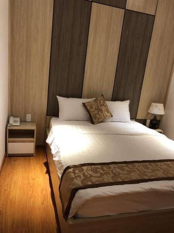 Habitación de hotel con cabecera de madera, cama con ropa blanca, mesita de noche con teléfono y lámpara en la mesita.