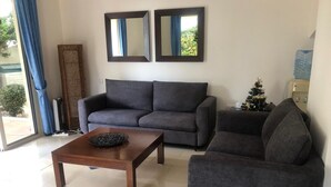 A living room with a grey sofa, a wooden coffee table, and a small armchair. There are two wall mirrors above the sofa. A Christmas tree is visible in the corner.