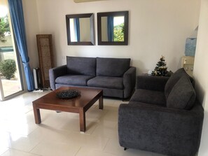 A living room with a grey sofa, a wooden coffee table, and a small armchair. There are two wall mirrors above the sofa. A Christmas tree is visible in the corner. - Private Villa with own pool 5 minutes drive to Coral Bay with 2 Sandy beaches (Paphos)