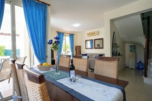 Un coin repas avec une table dressée pour deux personnes, une chaise en osier, un panier de fruits et une bouteille de vin.