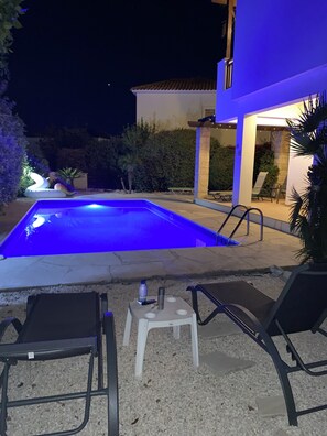 A swimming pool area with lounge chairs and a table, surrounded by a building with a tiled roof and a well-lit patio. - Private Villa with own pool 5 minutes drive to Coral Bay with 2 Sandy beaches (Paphos)