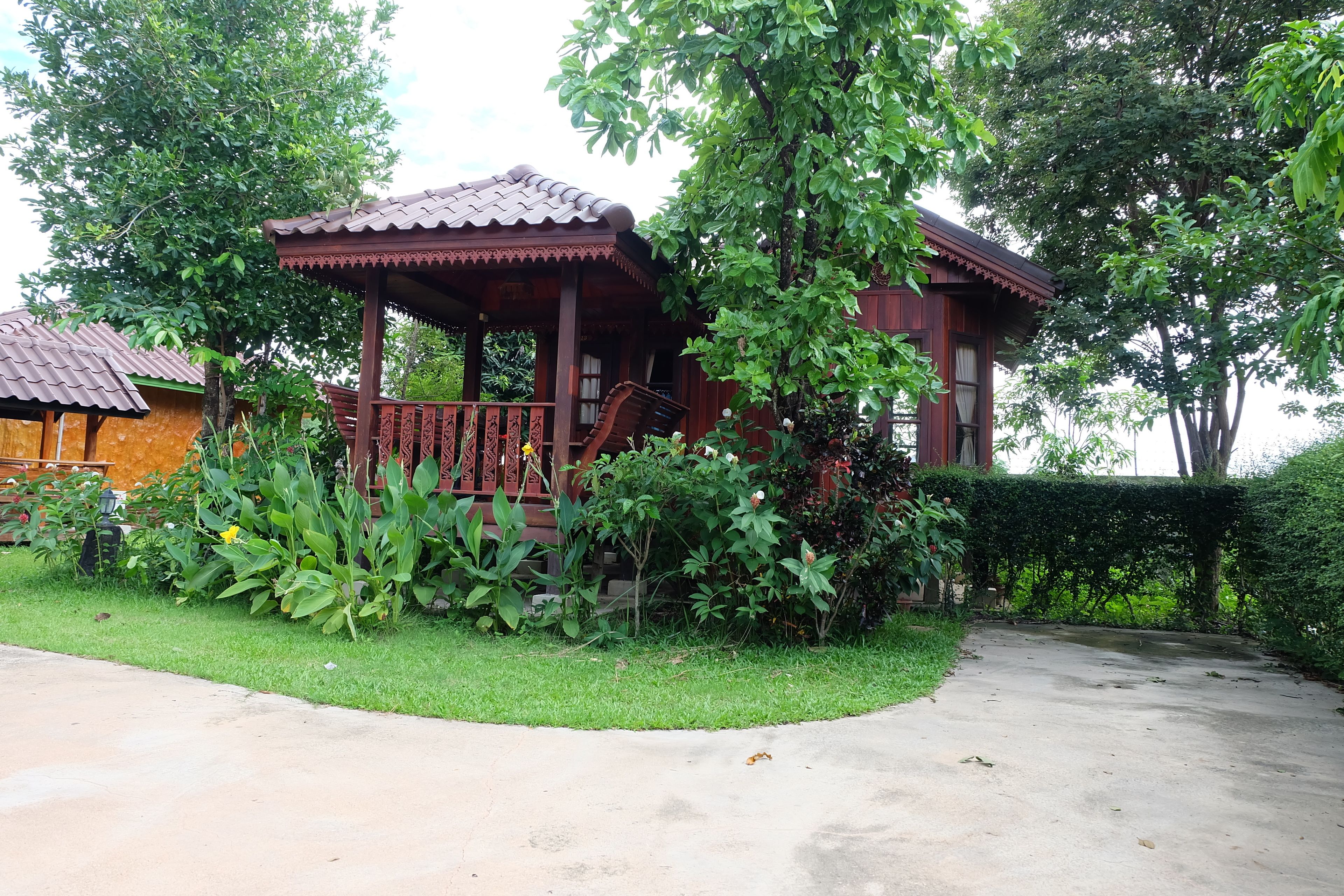 A wooden gazebo with a tiled roof, surrounded by lush greenery and a paved path.