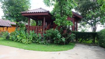 A wooden gazebo with a tiled roof, surrounded by lush greenery and a paved path.