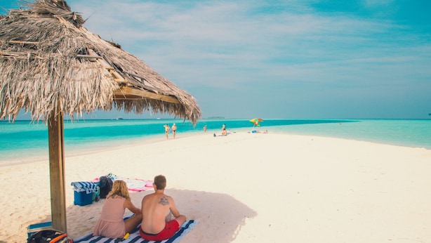 A beach scene with a thatched umbrella, a couple sitting on a striped towel, and a clear blue sea.