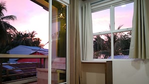 A room with a large window, wooden flooring, and a view of palm trees and buildings outside.